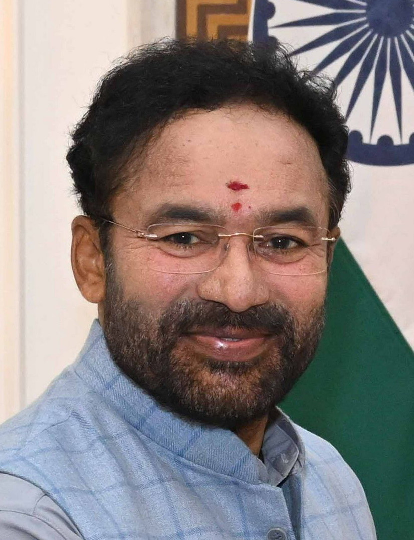Close-up portrait of G. Kishan Reddy in rimless glasses and a light blue checked waistcoat, with the Indian flag behind him