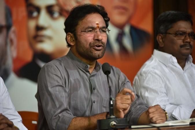 G. Kishan Reddy in a grey kurta and glasses speaking at a press conference table with a microphone, a colleague seated beside him