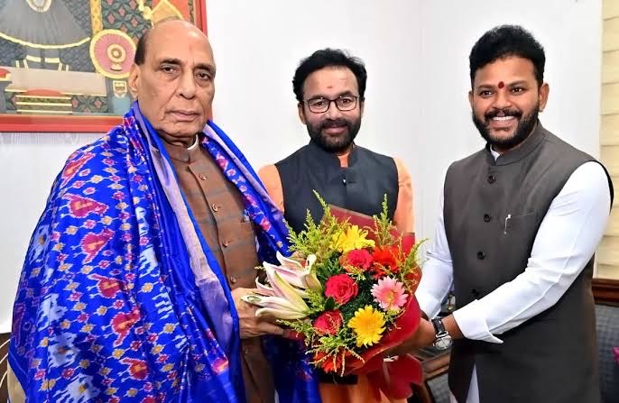 G. Kishan Reddy standing between two colleagues, one draped in a blue patterned shawl and holding a bouquet
