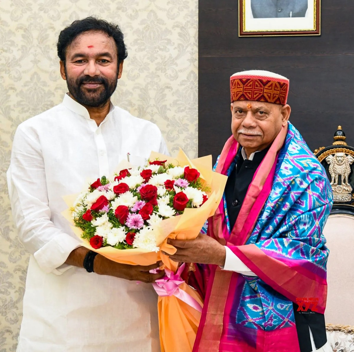 G. Kishan Reddy in white presenting a bouquet of red and white flowers to a guest wearing a traditional Himachali cap and a patterned shawl
