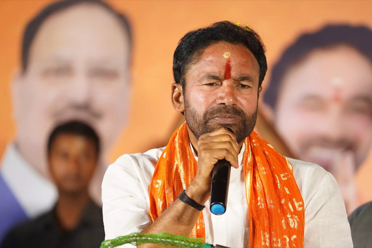 G. Kishan Reddy in a white kurta and saffron stole speaking into a handheld microphone at a public meeting, large portraits of party leaders behind him