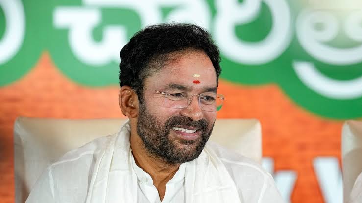 G. Kishan Reddy smiling broadly, wearing glasses and a white shawl, seated in front of a green and orange backdrop