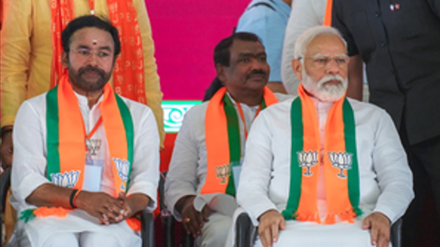 G. Kishan Reddy seated on stage beside Prime Minister Narendra Modi at a public meeting, both wearing saffron and green stoles