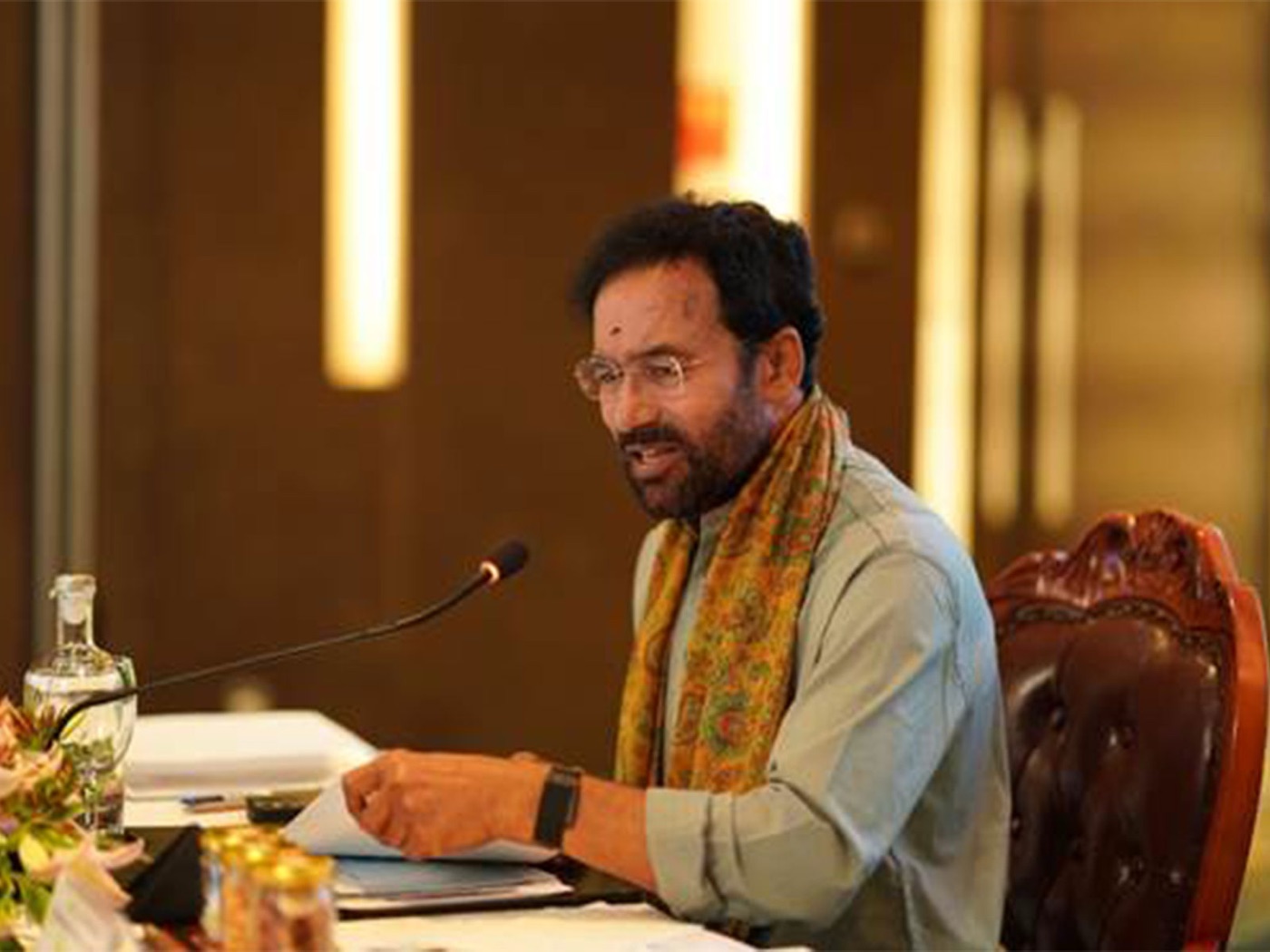 G. Kishan Reddy in glasses and a patterned stole speaking into a table microphone while chairing a meeting, papers in hand
