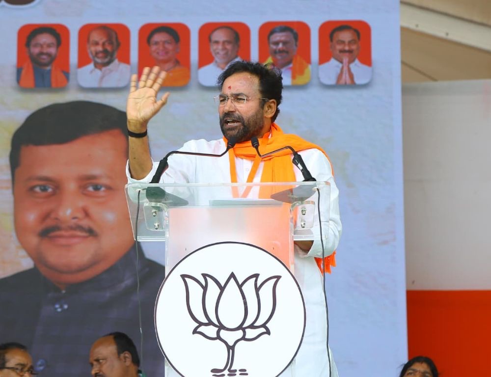 G. Kishan Reddy raising his hand while addressing a public meeting from a podium bearing the lotus symbol, with portraits of party leaders behind