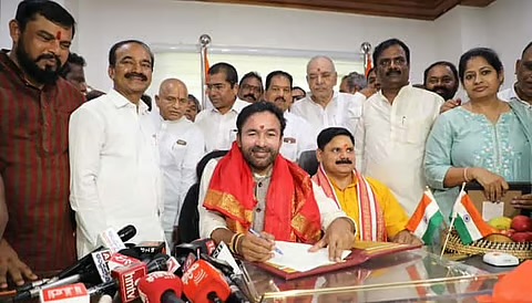 G. Kishan Reddy seated at a desk signing a file, surrounded by colleagues and officials, with news microphones in front