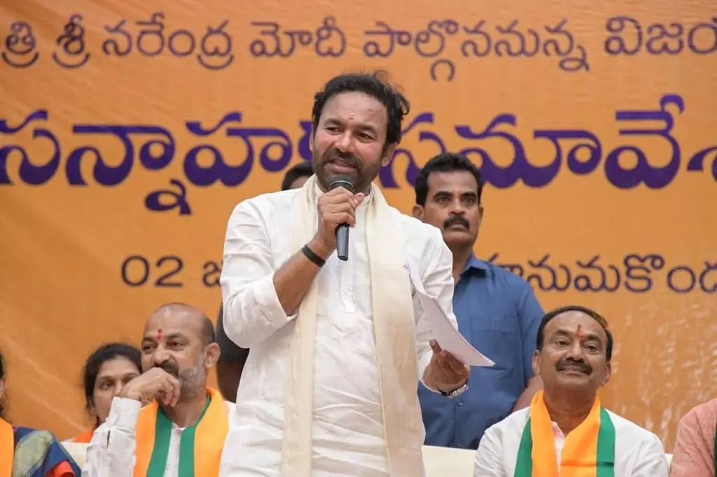 G. Kishan Reddy speaking into a microphone and holding notes at a meeting in Warangal, with a Telugu-language banner and seated colleagues