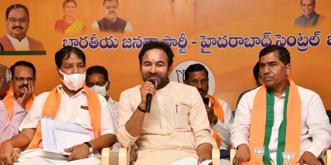 G. Kishan Reddy speaking into a microphone while seated among party office-bearers in saffron and green stoles at a BJP Hyderabad Central district meeting