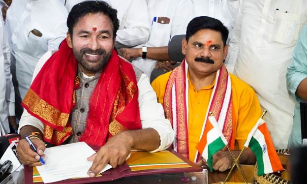 G. Kishan Reddy in a red and gold shawl smiling as he signs a document at a desk with miniature Indian flags, a colleague seated beside him