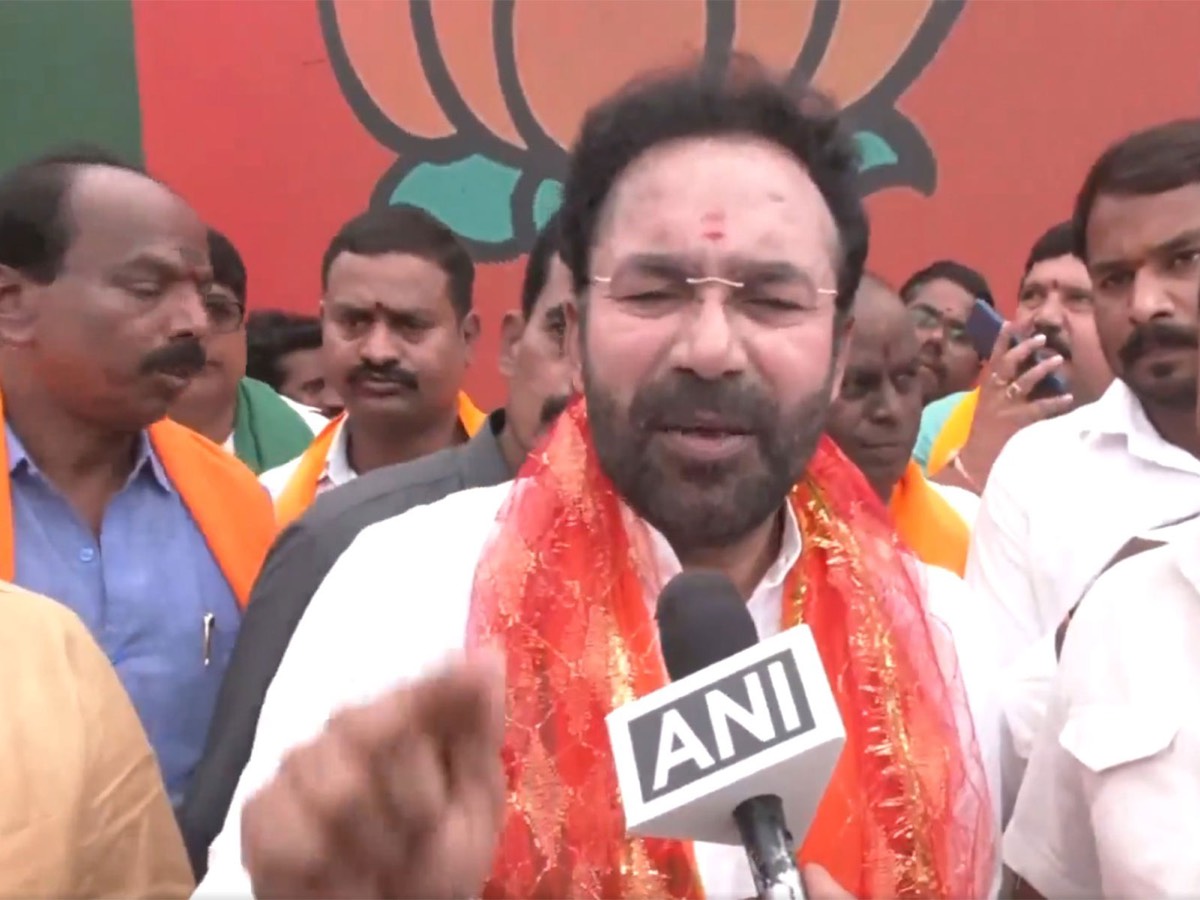 G. Kishan Reddy in a red and gold stole speaking to an ANI microphone, surrounded by party workers in saffron stoles in front of a lotus mural
