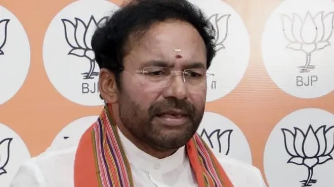 G. Kishan Reddy in glasses and a patterned stole speaking to reporters in front of a backdrop of lotus symbols at a party office