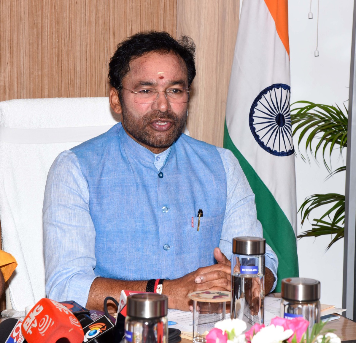 G. Kishan Reddy in a light blue waistcoat speaking at his desk with the Indian flag behind him, news microphones on the table