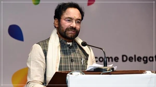 G. Kishan Reddy in a checked waistcoat and white shawl speaking at a lectern in front of a banner welcoming delegates